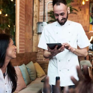 Portrait of happy bearded chef in uniform taking order himself via tablet. Guests are smiling and having fun with him in restaurant.